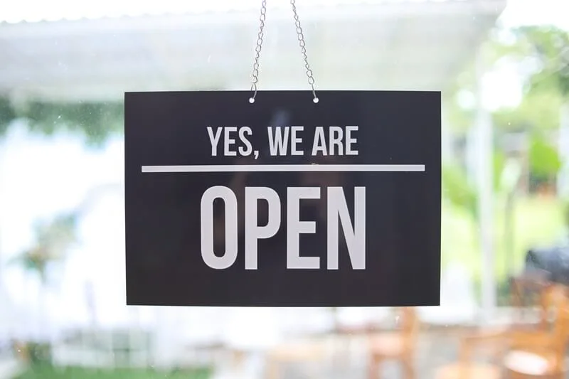 An image of a hanging door sign in black and white that says, "Yes, we are open."